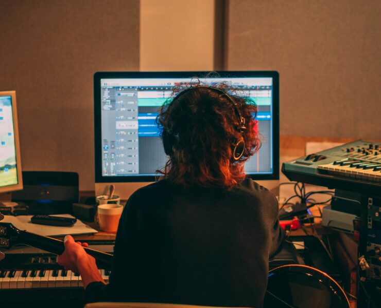Um produtor musical em seu estúdio caseiro, sentado em frente ao computador com um violão, compondo música.