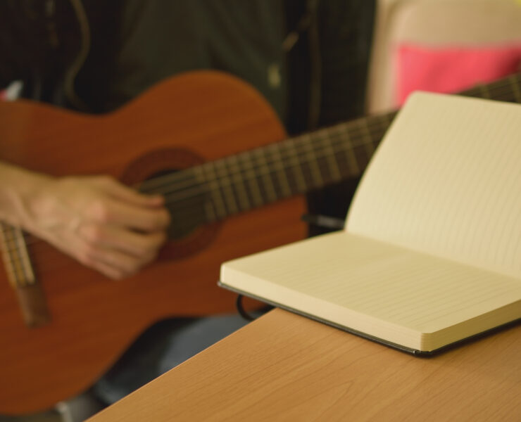 Ein Notizbuch liegt auf dem Schreibtisch, während im Hintergrund jemand Gitarre spielt.