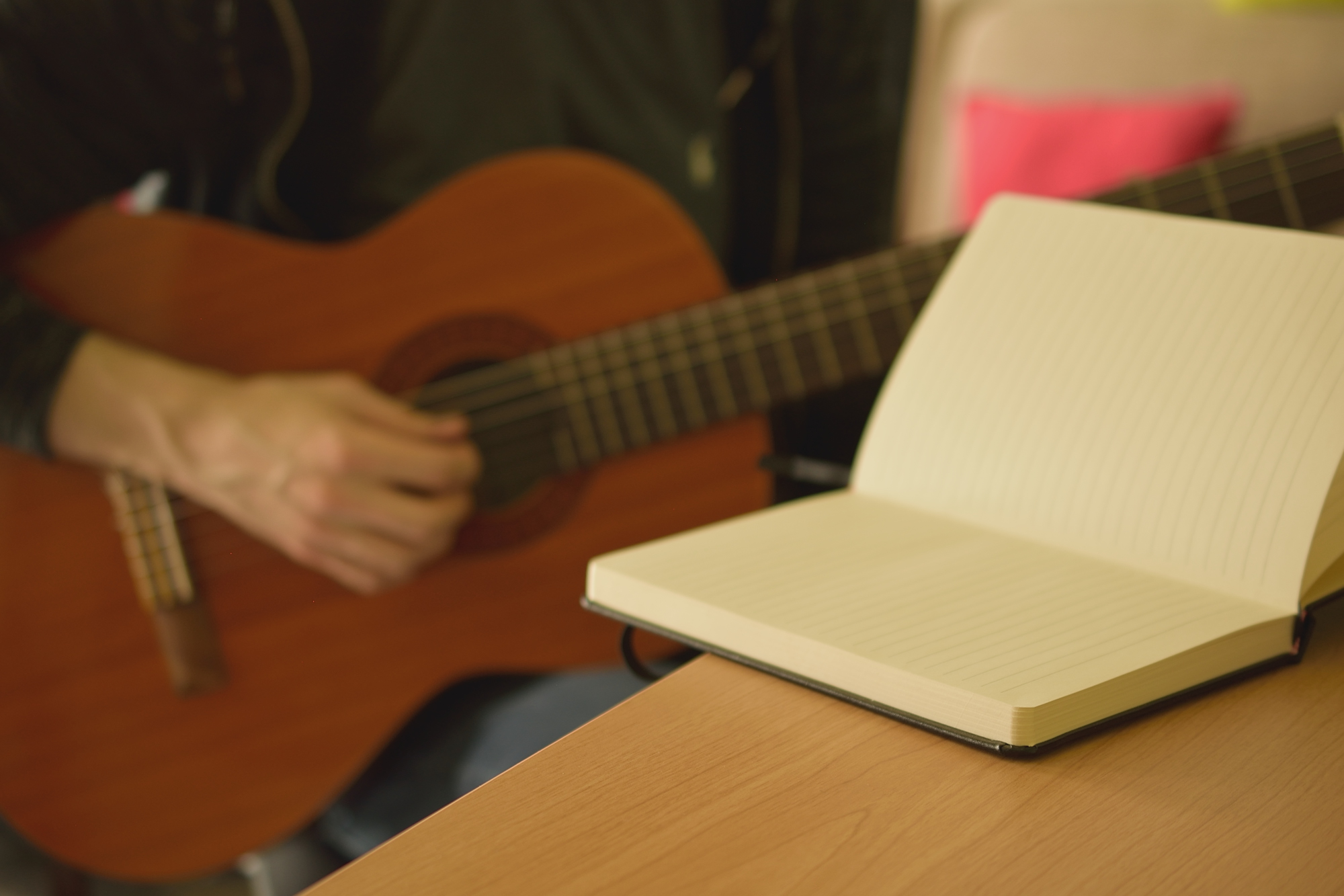 A notebook lies on the desk while someone is playing a guitar in the background.