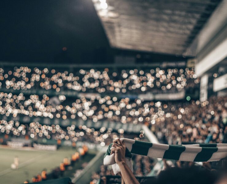 Un estadio lleno de gente cantando cánticos como ejemplo de música en eventos deportivos. Alguien sostiene una bufanda con los colores del equipo.