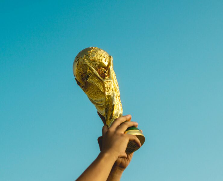 Manos levantando un trofeo de fútbol con el cielo azul de fondo.