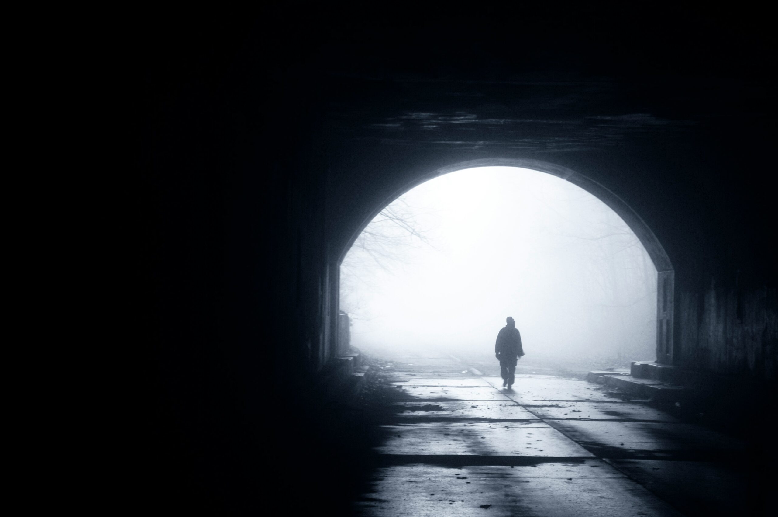 A cinematic and atmospheric photograph of a person walking out of a dark tunnel into a foggy forest road.